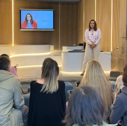 Formadora em palco durante evento corporativo, com público atento e ecrã de apresentação ao fundo
